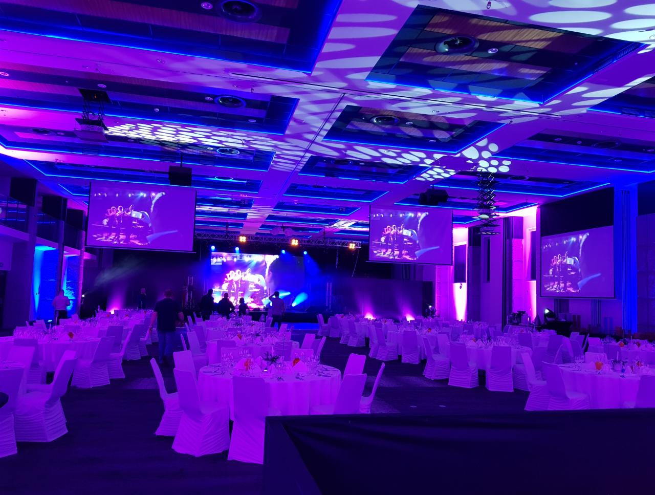 Sala bankietowa przygotowana na wydarzenie, stoły z białymi obrusami i krzesłami, oświetlenie sceniczne w odcieniach fioletu i niebieskiego, ekrany wyświetlające obraz na scenie w tle.
