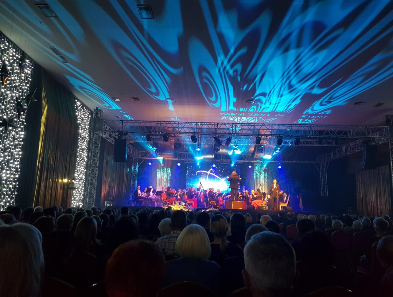 Widok na scenę z orkiestrą symfoniczną podczas koncertu, z widownią w tle i efektami świetlnymi na suficie.