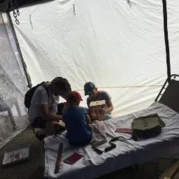 Wnętrze namiotu: mężczyzna i dwóch chłopców konstruują drewniany karmnik dla ptaków na łóżku polowym, obok leżą narzędzia i dokumenty.