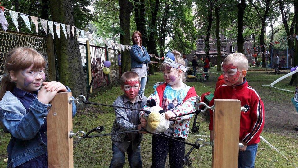 Dzieci z pomalowanymi twarzami bawią się w parku, używając drewnianej konstrukcji z elastycznymi linami i pluszowej zabawki Angry Birds.