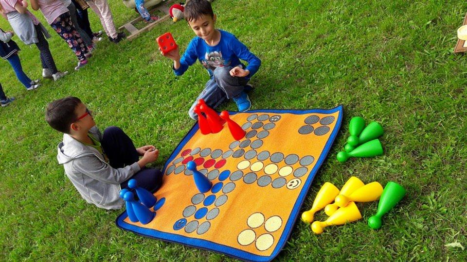 Dzieci grają w plenerową wersję gry planszowej Chińczyk na trawie w Rudzie Śląskiej, używając dużych pionków i kostki.