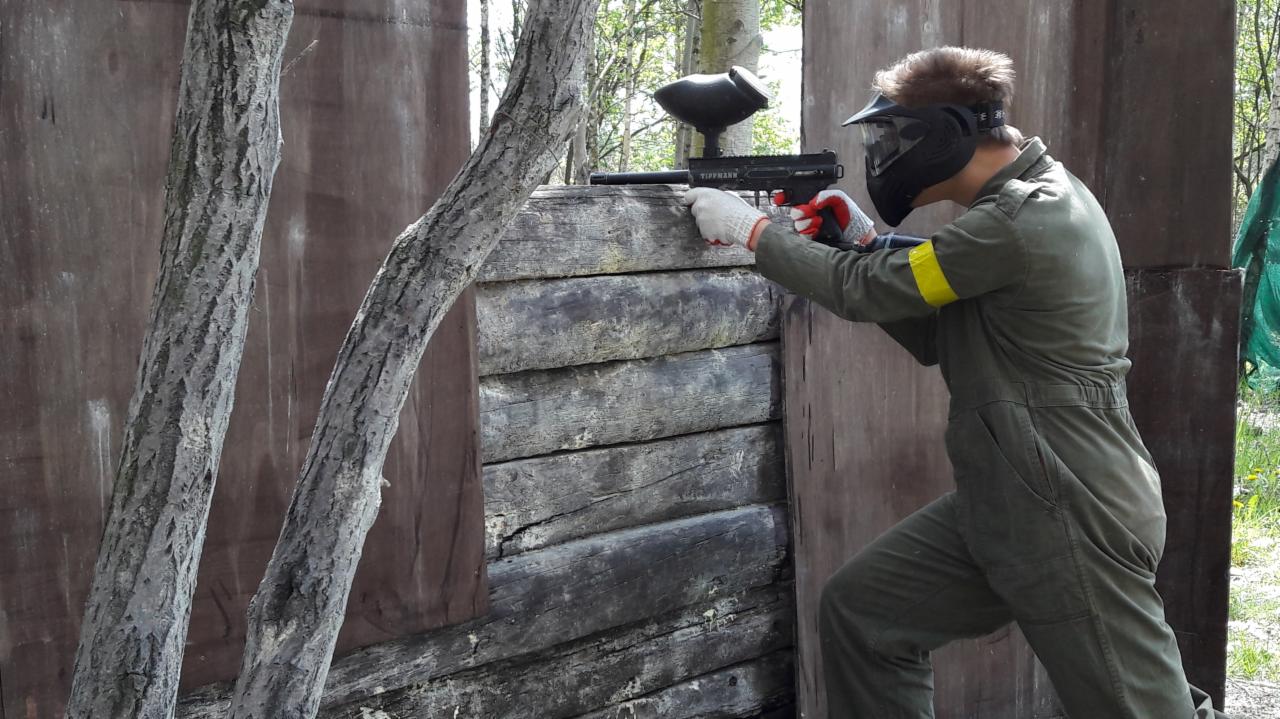 Osoba w kombinezonie i masce ochronnej, celująca markerem paintballowym zza drewnianej barykady z widocznym lasem w tle.