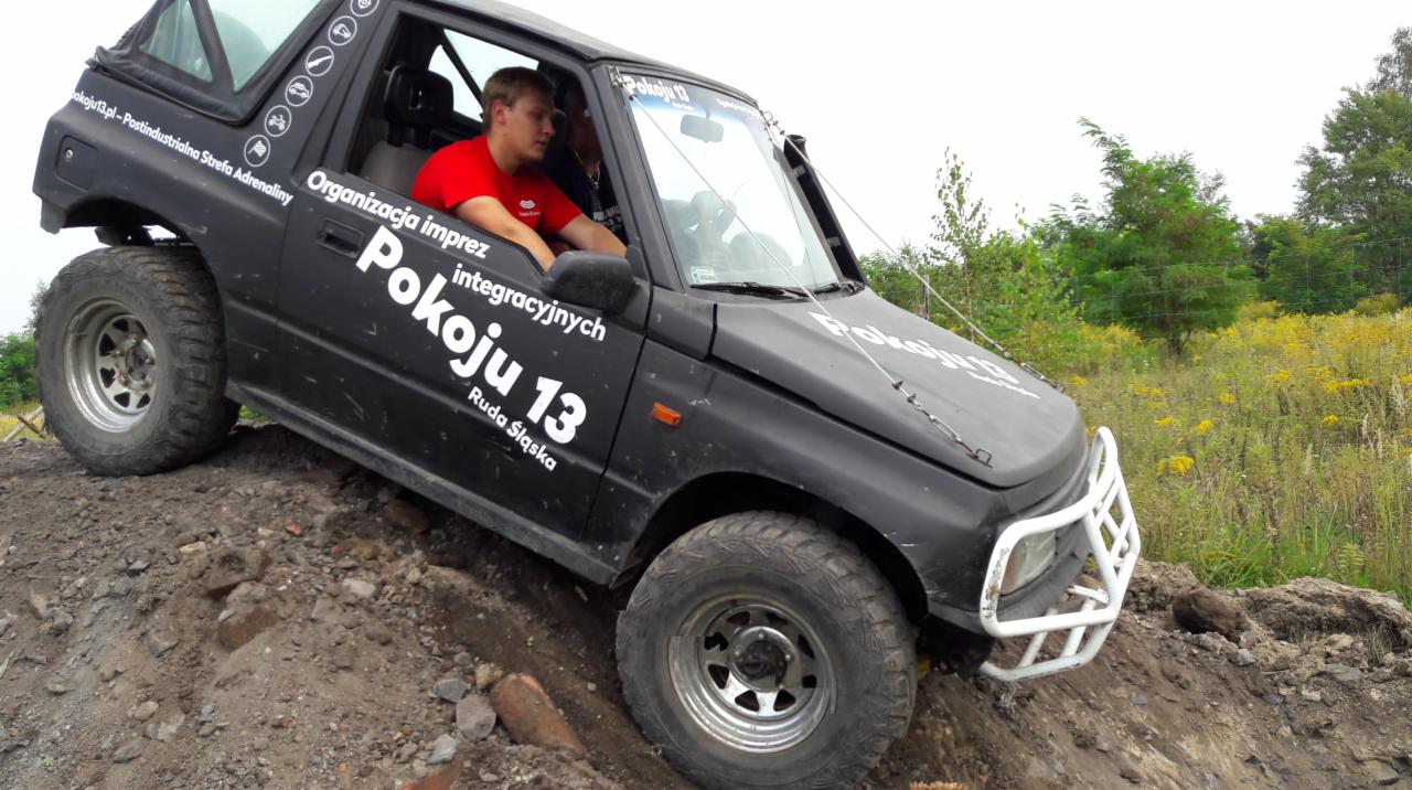Czarny samochód terenowy z napisem 'Organizacja imprez Pokoju 13 Ruda Śląska' pokonuje terenowy tor przeszkód, widoczni kierowcy wewnątrz.