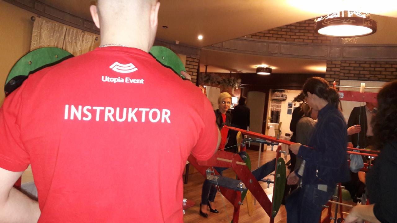 Instruktor w czerwonej koszulce z napisem 'Utopia Event' nadzoruje uczestników podczas budowy konstrukcji z drewnianych elementów podczas imprezy integracyjnej.