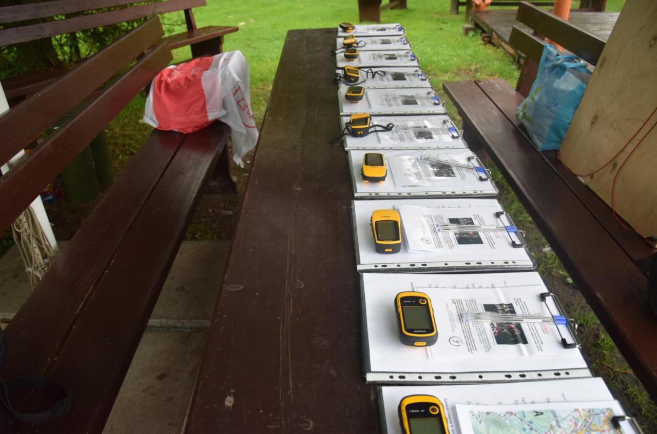 Przygotowane stanowiska do gry terenowej: żółte odbiorniki GPS, mapy i długopisy na drewnianym stole z ławkami w plenerze.