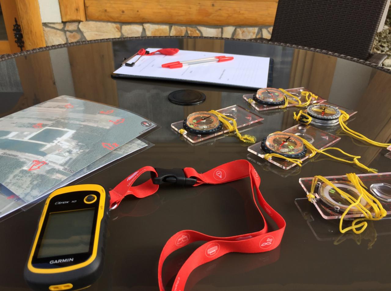 Przygotowanie do gry terenowej: mapa z zaznaczonymi punktami, kompas z żółtym sznurkiem, GPS Garmin Etrex 10 i notatnik z długopisem na szklanym stole.