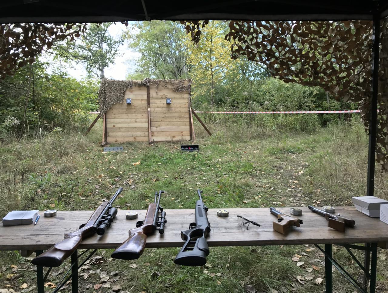 Stanowisko strzeleckie z kilkoma karabinkami pneumatycznymi na drewnianym stole, tarcza z dwoma otworami w tle, widok z zadaszonego miejsca.