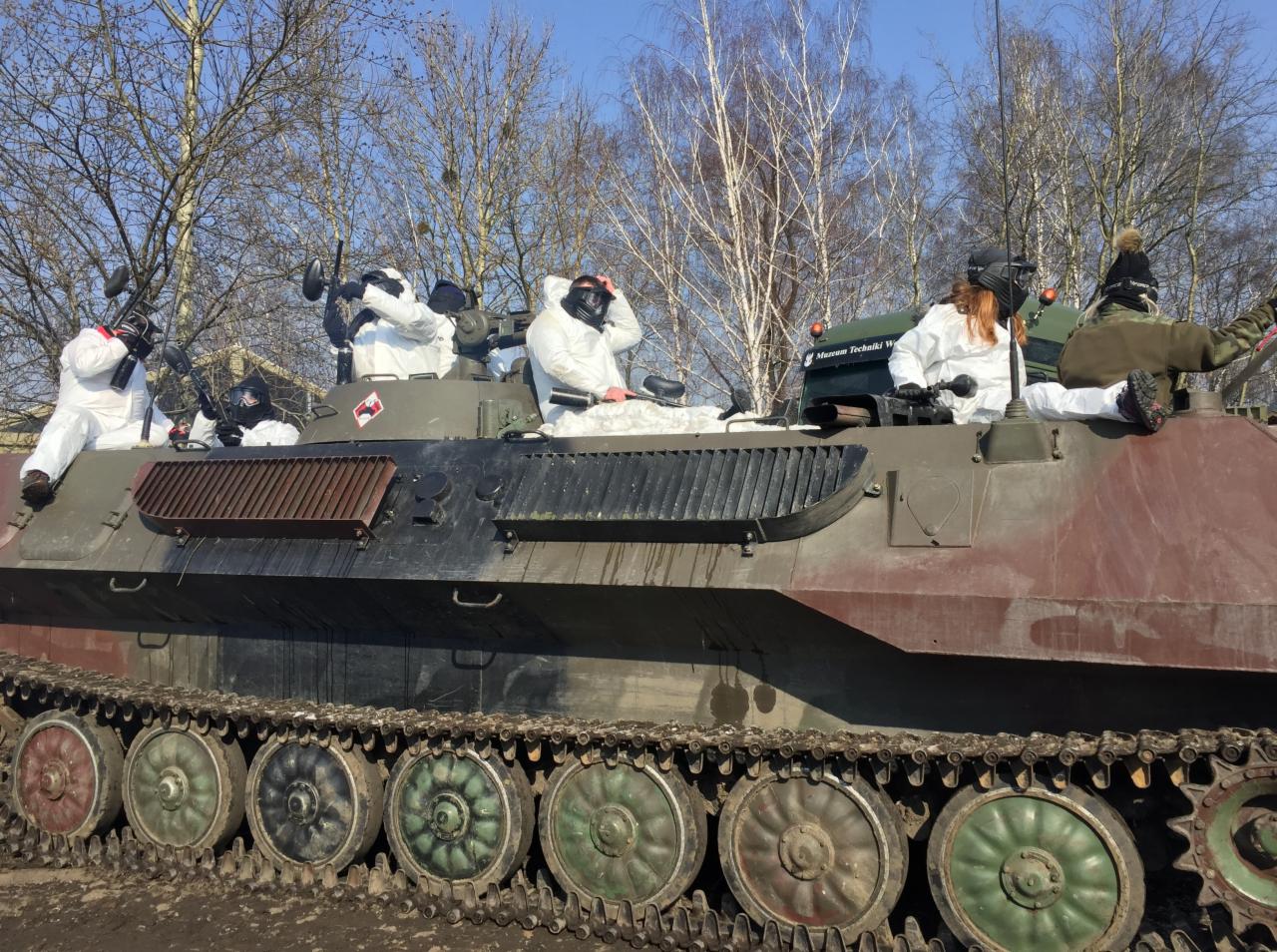 Grupa osób w białych kombinezonach i maskach na wojskowym pojeździe opancerzonym; prawdopodobnie gra paintball lub laser tag w plenerze.