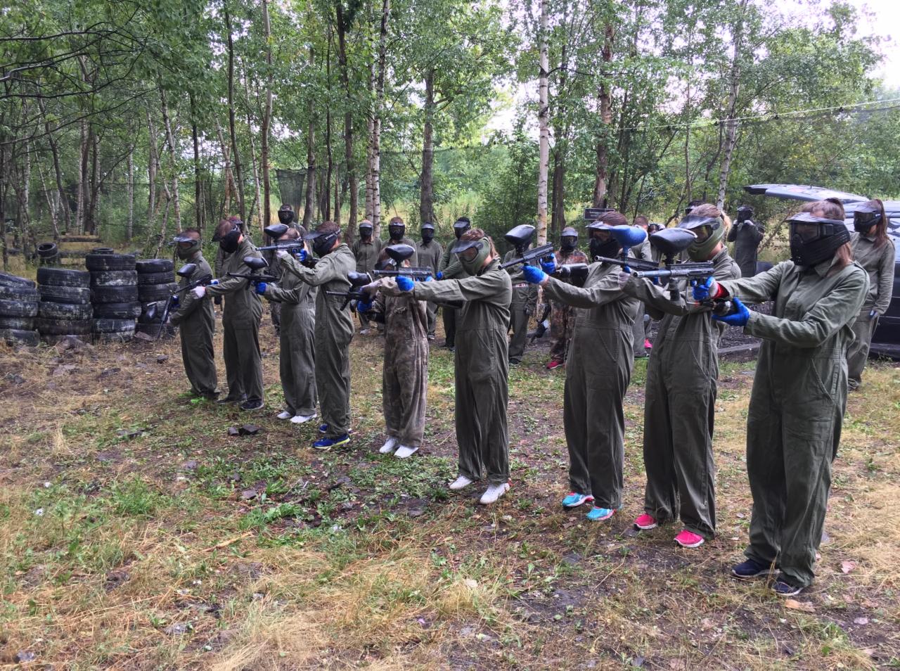Grupa osób w kombinezonach i maskach ochronnych celuje z markerów paintballowych na zewnątrz, w tle las i stos opon.