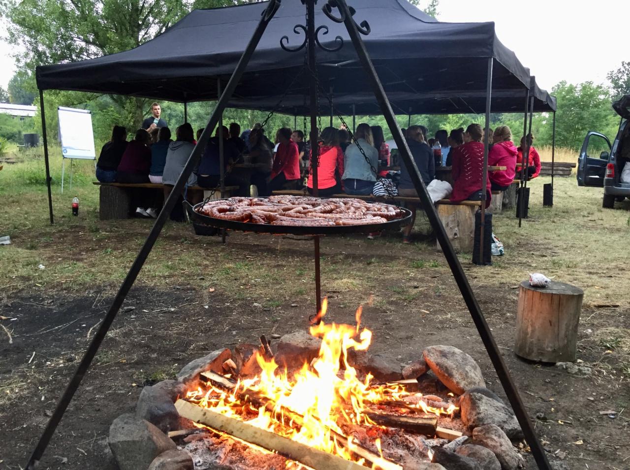 Ognisko z rusztem pełnym kiełbas, w tle grupa osób siedząca pod namiotem podczas spotkania na świeżym powietrzu.