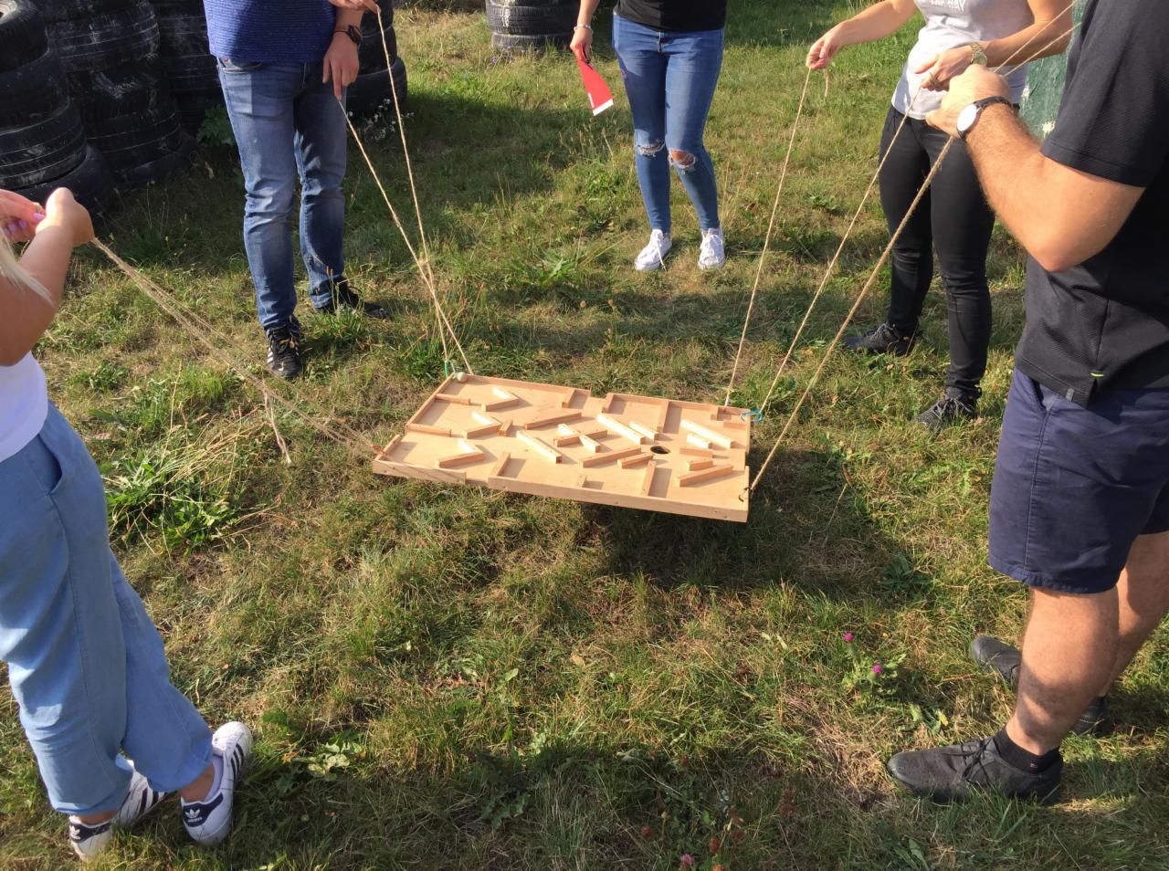 Grupa osób grająca w grę zręcznościową na trawie, polegającą na prowadzeniu kulki przez labirynt za pomocą sznurków.