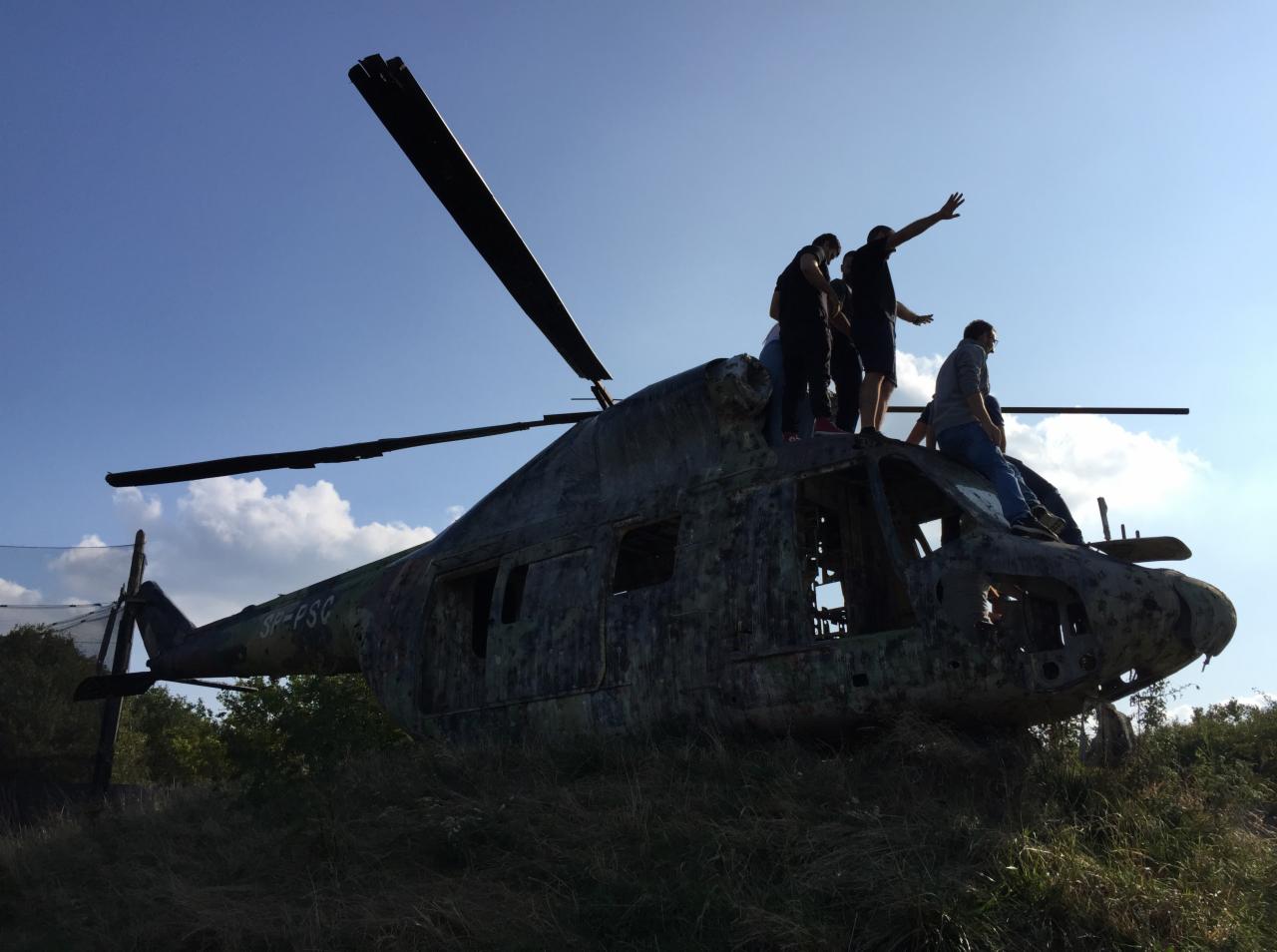 Grupa osób stoi na zniszczonym helikopterze na tle błękitnego nieba z chmurami. Helikopter pokryty jest farbą, a trawa rośnie wokół niego.