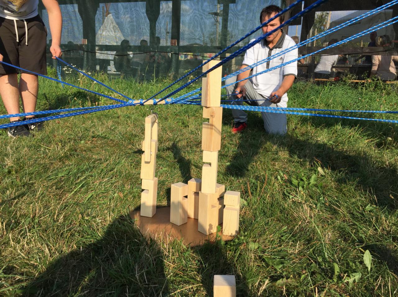 Gra zespołowa na trawie: uczestnicy za pomocą sznurków sterują drewnianymi klockami, próbując zbudować wieżę.