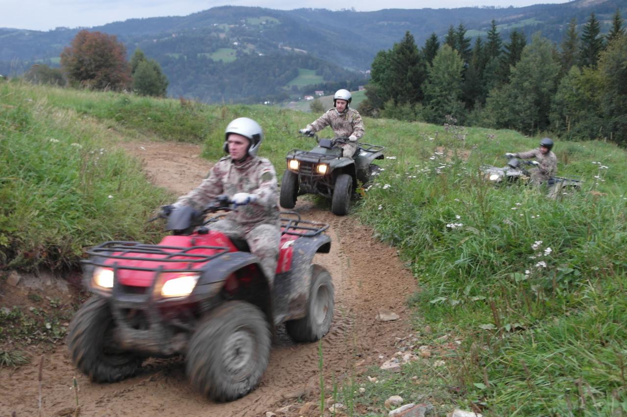 Trzech mężczyzn w kaskach i kamuflażu jedzie quadami po błotnistej, polnej drodze na tle gór i lasów.