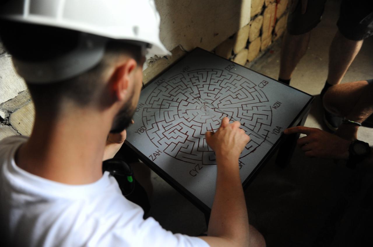 Mężczyzna w kasku wskazuje palcem na labirynt z kodami na kwadratowym stole; widoczni uczestnicy zabawy.