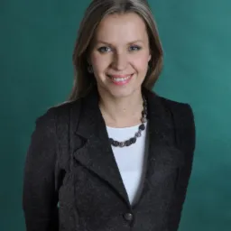 Portret uśmiechniętej kobiety w ciemnoszarym żakiecie i białej bluzce, z naszyjnikiem z ciemnych kamieni na tle ciemnozielonego tła.