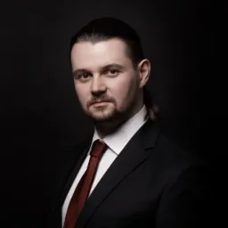 Portret mężczyzny w czarnym garniturze i bordowym krawacie na ciemnym tle, z włosami związanymi w niski kucyk.