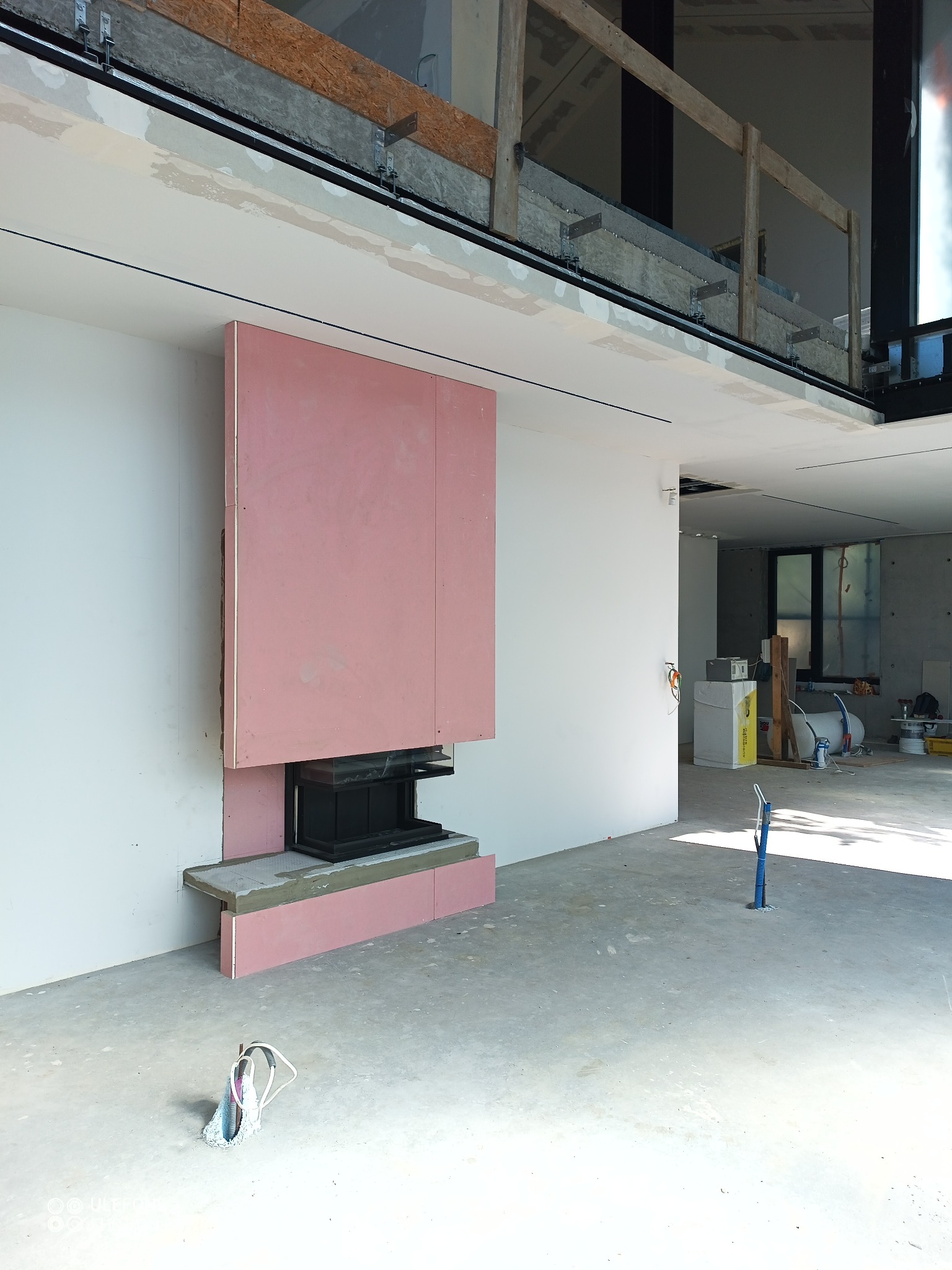 Wnętrze w trakcie remontu z zamontowanym wkładem kominkowym obudowanym płytami w kolorze różowym, widoczne instalacje elektryczne i hydrauliczne w podłodze.