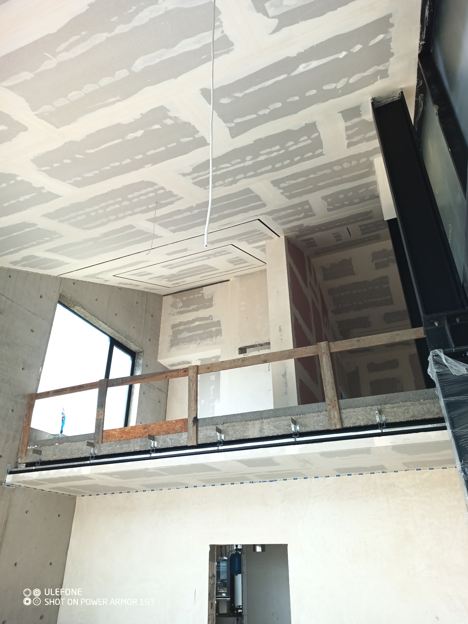 Wnętrze w trakcie remontu: widok na sufit z zamontowanymi płytami i widocznymi łączeniami, częściowo zaszpachlowanymi, z elementami konstrukcyjnymi budynku i prowizoryczną barierką zabezpieczającą.