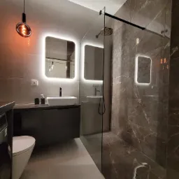 Nowoczesna łazienka z szarymi płytkami imitującymi marmur, podświetlanym lustrem, wiszącą lampą z żarówką Edisona i kabiną prysznicową typu walk-in z deszczownicą.