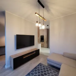 Nowoczesny salon z szarą sofą narożną, czarnym telewizorem na drewnianej szafce RTV i designerską lampą wiszącą z trzema żarówkami w klatkach, widok na kuchnię z drewnianymi szafkami.