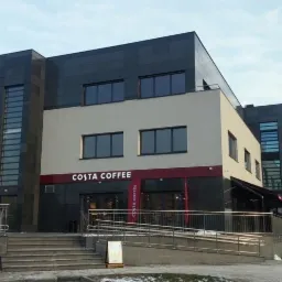 Nowoczesny budynek kawiarni Costa Coffee z grafitową elewacją z płyt, jasną częścią środkową i dużymi oknami, widok z zewnątrz w pochmurny dzień.