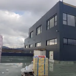 Budynek przemysłowy z ciemną elewacją z płyt warstwowych w trakcie montażu, widoczne materiały izolacyjne na dachu, pochmurne niebo w tle.