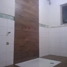 Montaż kabiny prysznicowej z panelami imitującymi drewno na ścianie w łazience z białymi płytkami.