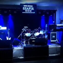 Scena z instrumentami muzycznymi i oświetleniem w kolorze niebieskim, widoczny zestaw perkusyjny, gitary, keyboardy oraz logo zespołu 'Szafir' na telebimie w tle.