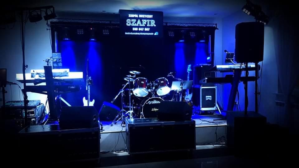 Scena z instrumentami muzycznymi i oświetleniem w kolorze niebieskim, widoczny zestaw perkusyjny, gitary, keyboardy oraz logo zespołu 'Szafir' na telebimie w tle.