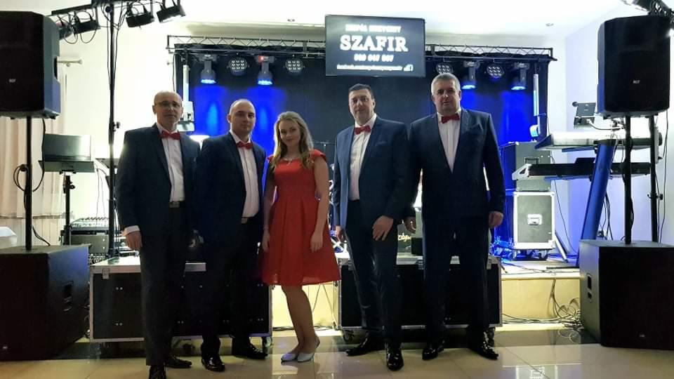 Pięcioosobowy skład muzyczny w garniturach i czerwonych muchach pozuje na tle sceny z oświetleniem i nagłośnieniem, widoczny baner z nazwą zespołu.