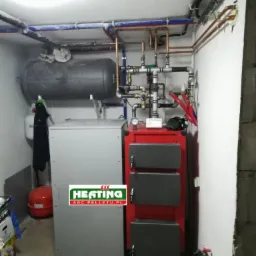 Kotłownia z piecem na paliwo stałe, zbiornikiem akumulacyjnym i instalacją grzewczą, widoczne rury miedziane i plastikowe, logo firmy na piecu.