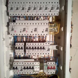 Rozdzielnica elektryczna z zainstalowanymi bezpiecznikami, przewodami w różnych kolorach oraz listwami zaciskowymi, widoczna na tle ściany z surową cegłą.