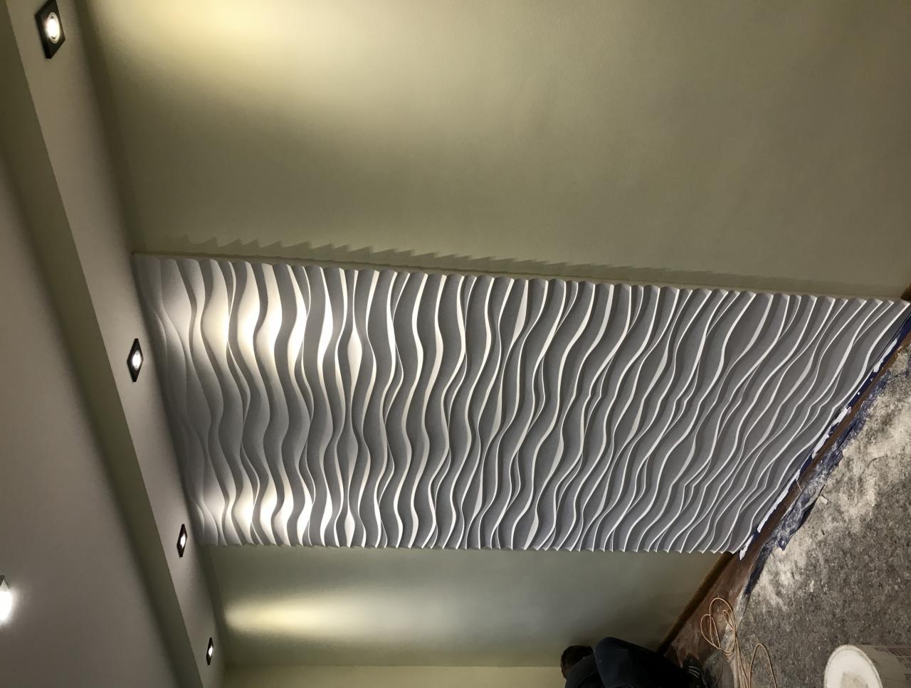 Montaż dekoracyjnych paneli ściennych 3D o falistym wzorze, widoczne oświetlenie punktowe i częściowo zakryta osoba podczas pracy.