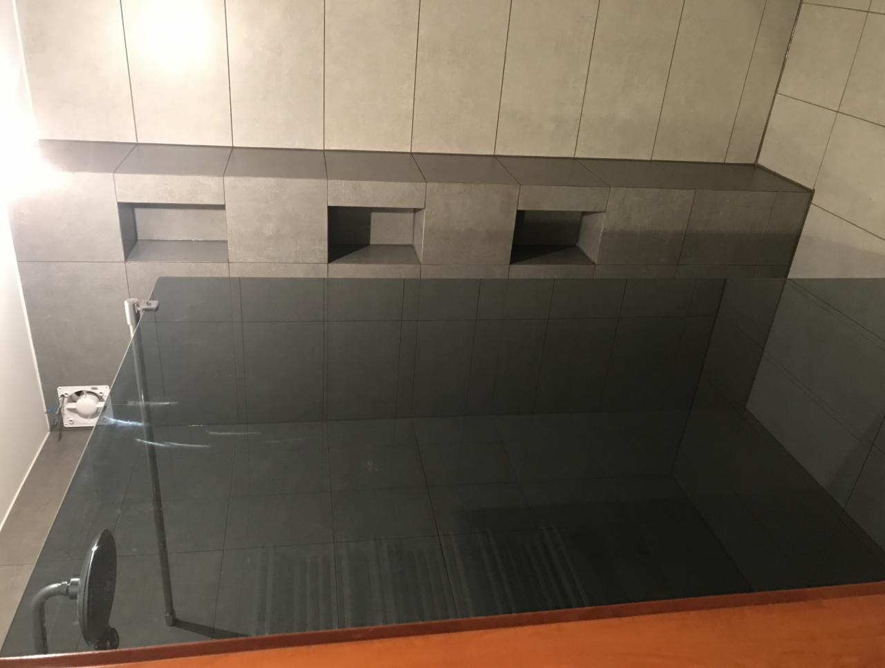 Nowoczesna kabina prysznicowa z szarymi płytkami, wbudowanymi półkami w ścianie i szklanymi drzwiami, widoczne mocowania i odpływ.