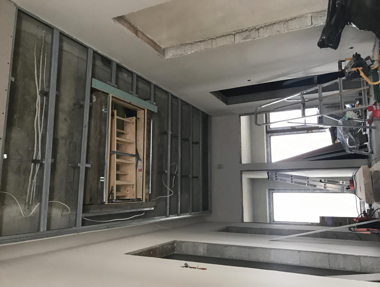 Montaż metalowej konstrukcji pod płytę gipsową, widoczne kable elektryczne, otwór na schody strychowe z drewnianą ramą i półkami, oraz drabina i rusztowanie w tle.