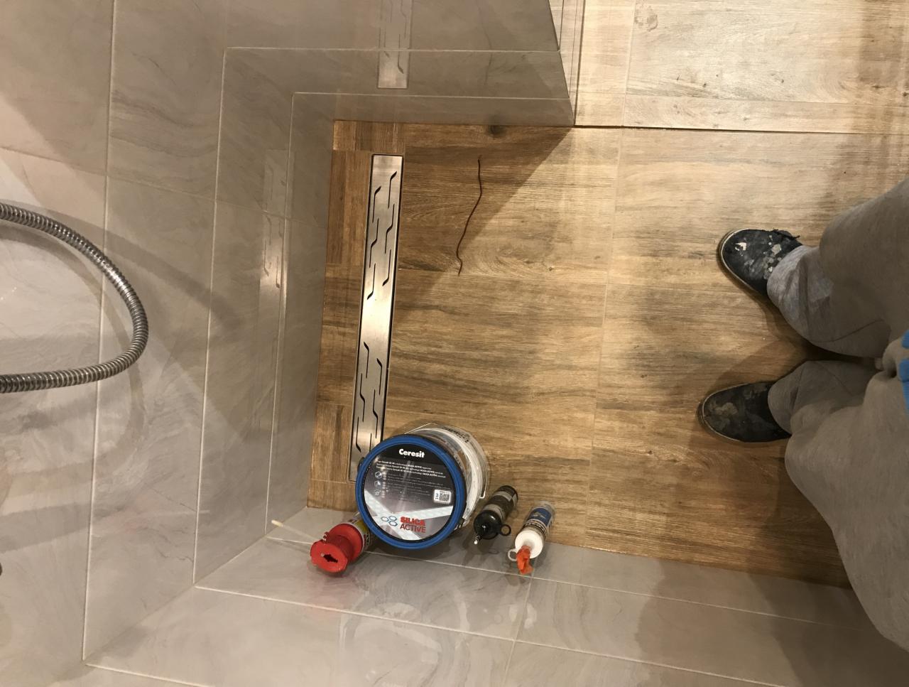 Montaż odpływu liniowego w kabinie prysznicowej z płytkami imitującymi drewno, widoczne materiały uszczelniające Cerisit i fragment nóg osoby wykonującej prace.