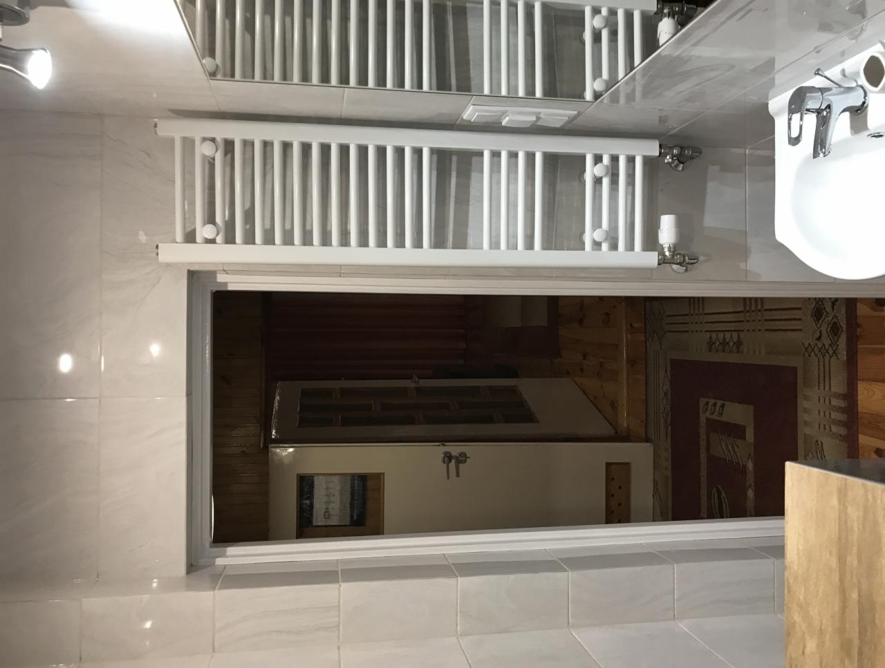 Biała łazienka z grzejnikiem drabinkowym, umywalką i widokiem na pokój z drewnianą podłogą i dywanem przez otwarte drzwi.