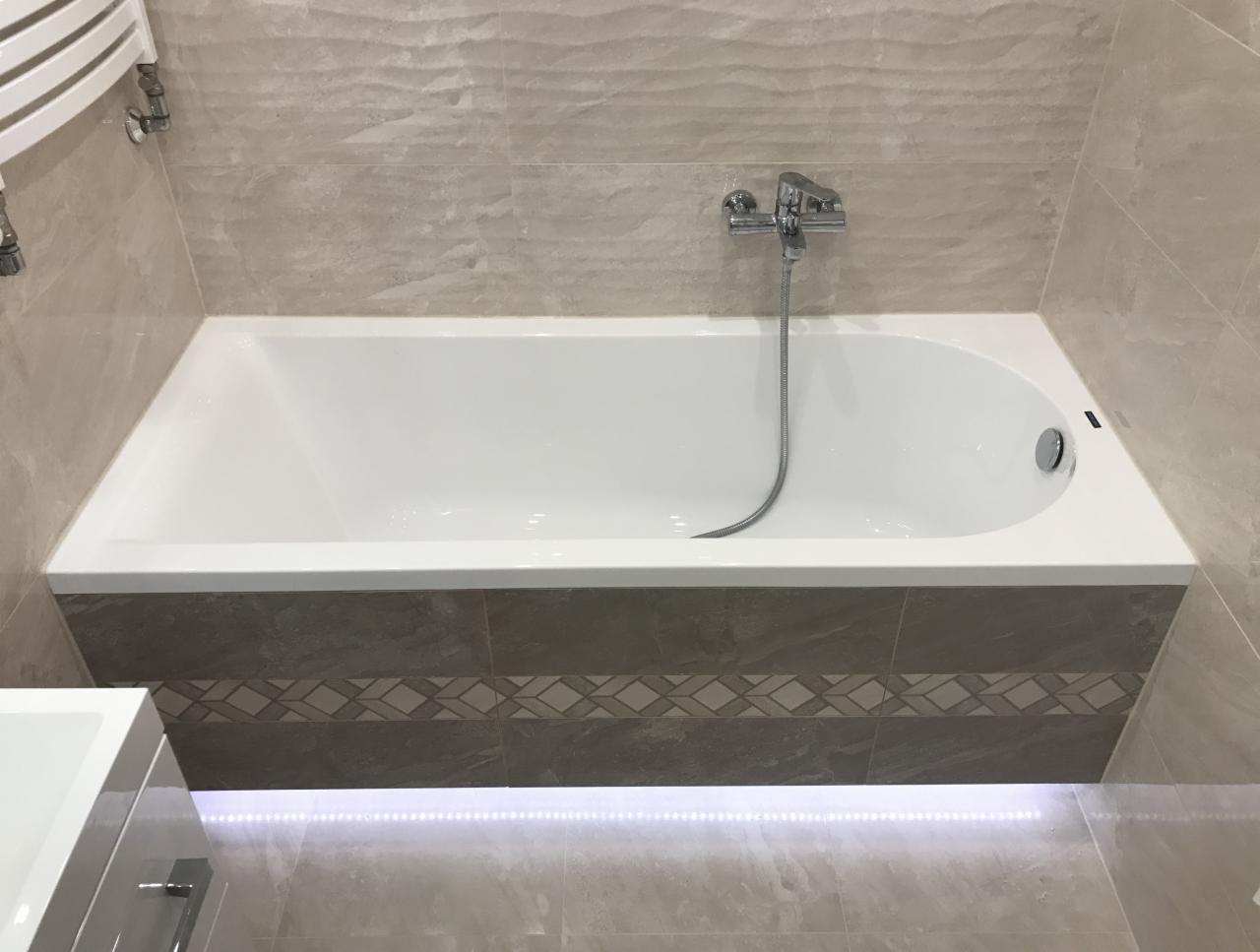 Biała wanna z chromowaną baterią i szarą obudową z dekoracyjnym pasem, beżowe płytki na ścianie, oświetlenie LED pod wanną.