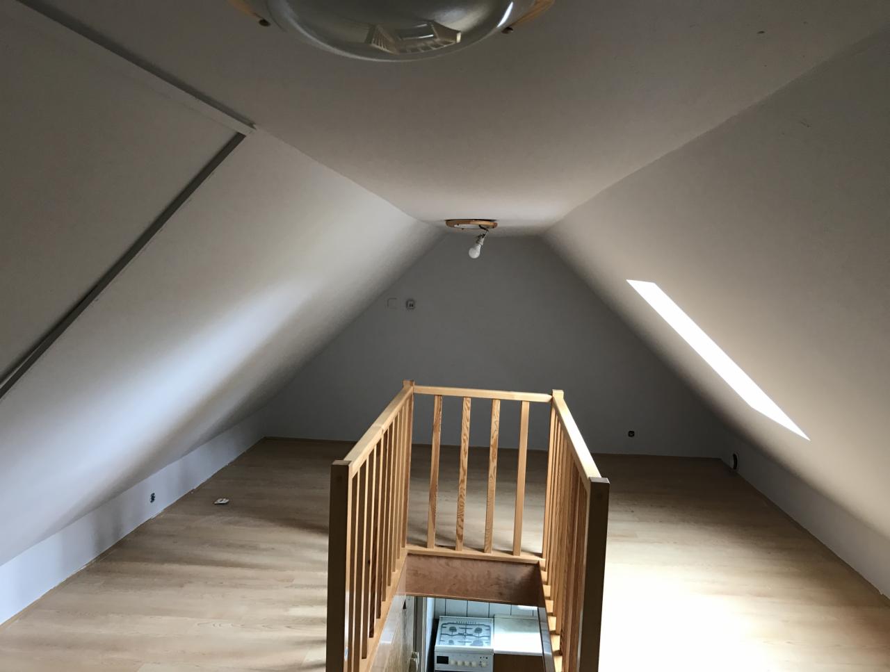 Adaptacja poddasza: widok na przestrzeń z drewnianą balustradą, skosy ścian pomalowane na biało, podłoga wyłożona panelami, widoczny fragment kuchni na niższym poziomie.