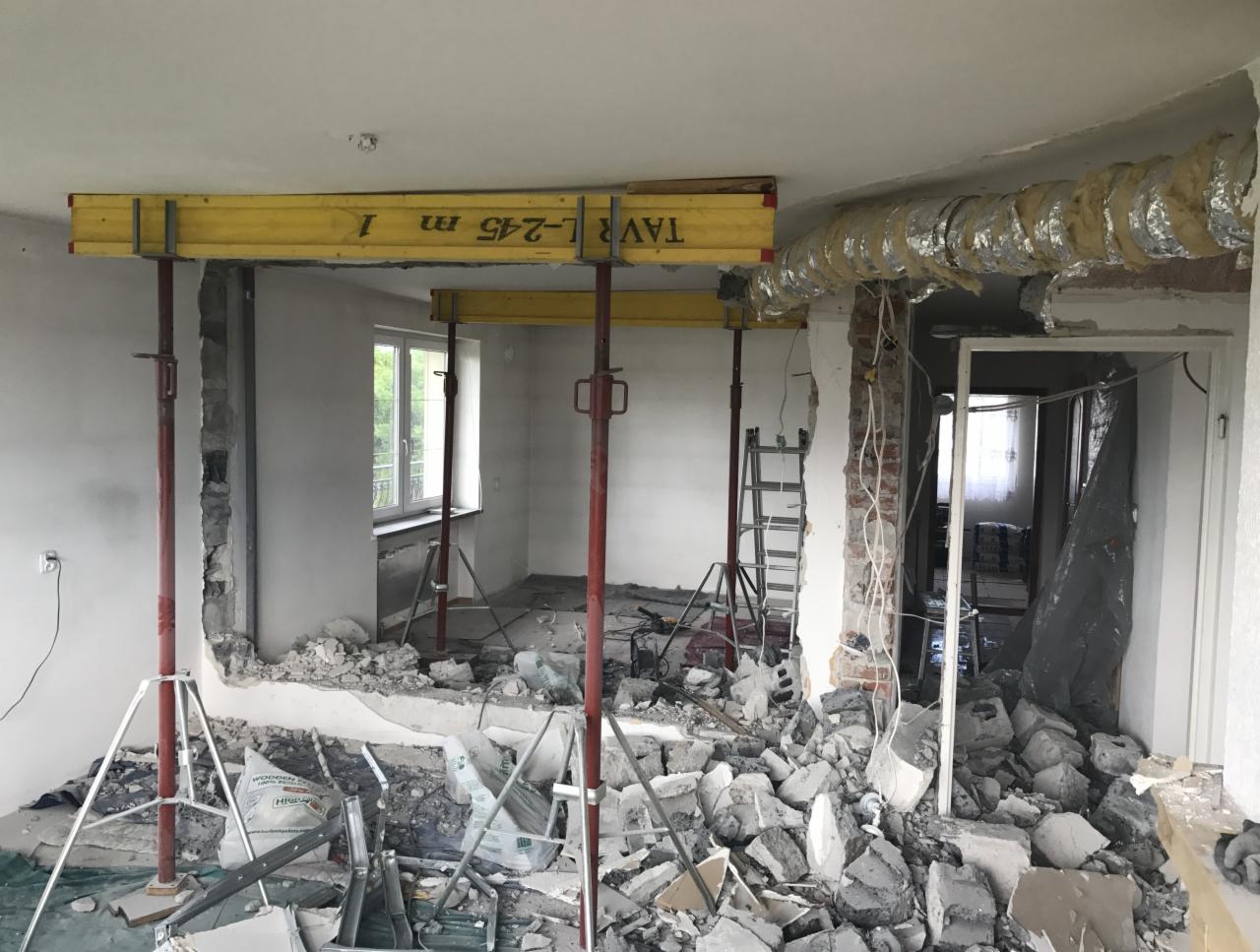 Wnętrze w trakcie demontażu ściany działowej, widoczne podpory stropu, gruz, odsłonięte przewody elektryczne i częściowo zdemontowana wentylacja.