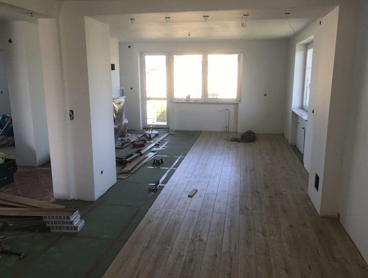 Wnętrze w trakcie układania podłogi z paneli drewnopodobnych; widoczne narzędzia, materiały i fragmenty nieukończonej podłogi na zielonej macie izolacyjnej.