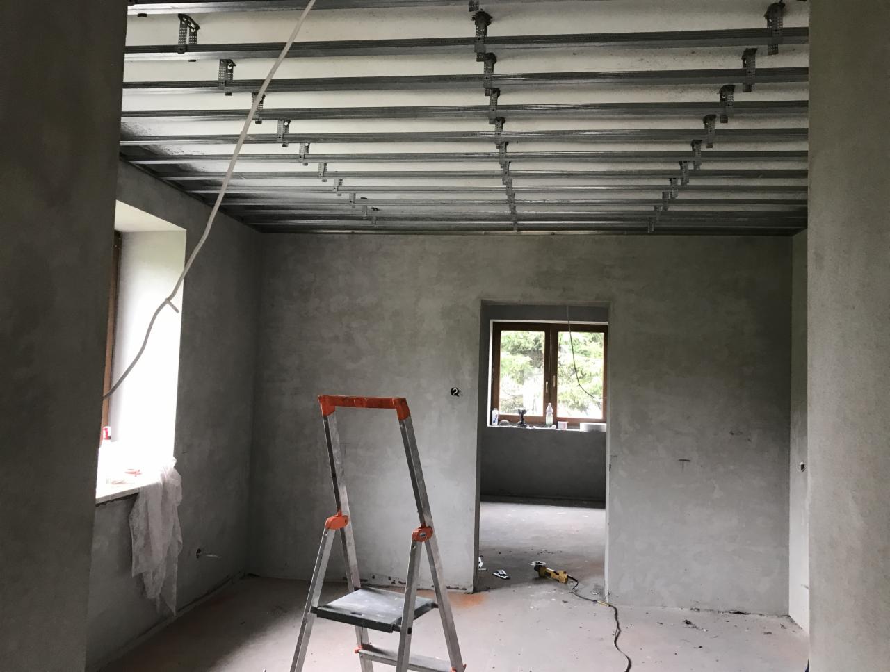 Surowy stan wnętrza z zamontowaną metalową konstrukcją podwieszanego sufitu, widoczna drabina i narzędzia, ściany pokryte szarym tynkiem, okno z widokiem na zieleń.