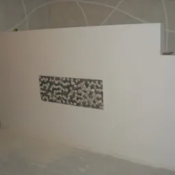 Świeżo wykończona ściana z prostokątną wnęką ozdobioną drobnymi kamieniami, widoczne ślady prac wykończeniowych na podłodze, fragment rusztowania z lewej strony.