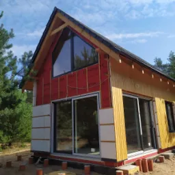 Dom w trakcie ocieplania z widocznymi panelami izolacyjnymi, membraną wiatroizolacyjną i drewnianą elewacją, okna zamontowane, teren wokół domu piaszczysty z elementami konstrukcyjnymi.