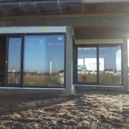 Montaż dużych, przesuwnych okien z czarnymi ramami w surowym stanie budynku, widoczne bloczki fundamentowe i przygotowany teren.