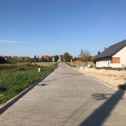 Nowa, szara nawierzchnia z kostki brukowej prowadząca przez osiedle domów jednorodzinnych w słoneczny dzień. W tle widoczne domy w różnej fazie budowy.