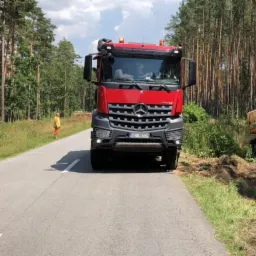 Czerwona ciężarówka Mercedes i żółta koparka Liebherr podczas prac ziemnych przy drodze w otoczeniu lasu iglastego, widoczny pracownik w pomarańczowej kamizelce.