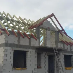 Surowy stan domu z białego pustaka z drewnianą konstrukcją dachu, widoczna drabina i materiały budowlane. Etap budowy, widok z dołu na tle pochmurnego nieba.