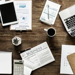 Biurko z laptopem, tabletem, kalkulatorem, gazetą, kubkiem kawy i dokumentami finansowymi, z góry.